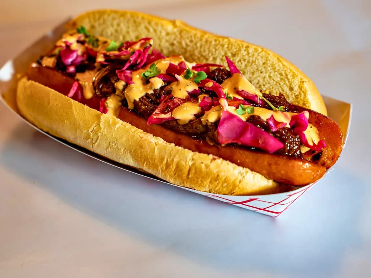 The Dam Dog — a split, griddled dog under pulled BBQ brisket, pink pickled cabbage, hot sauce aioli, and micro greens, in a paper boat.