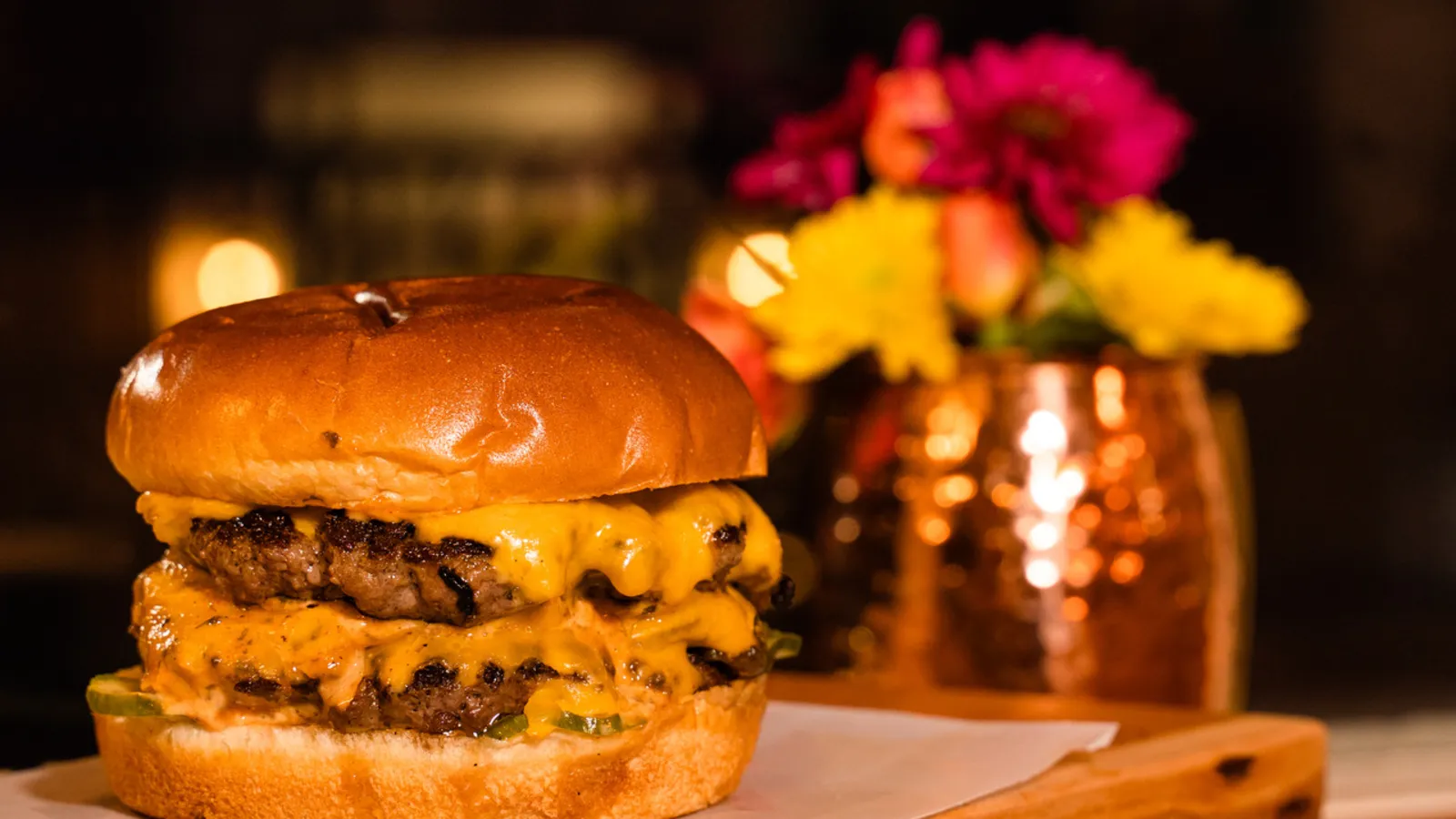 The Burgerly cheeseburger on a brioche bun with savory onions and house pickles, photographed warm on a live-edge wood board at The Burgerly in New Hope, PA.