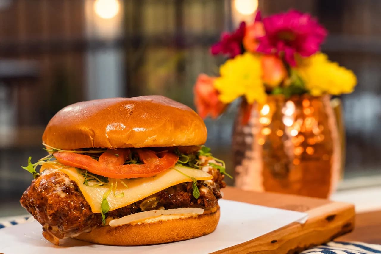 Chicken Flew Southwest — fried marinated chicken breast with muenster, fresh tomato, micro cilantro, and spicy serrano pepper honey ranch on a brioche bun, photographed warm with a copper mule and floral arrangement at The Burgerly.