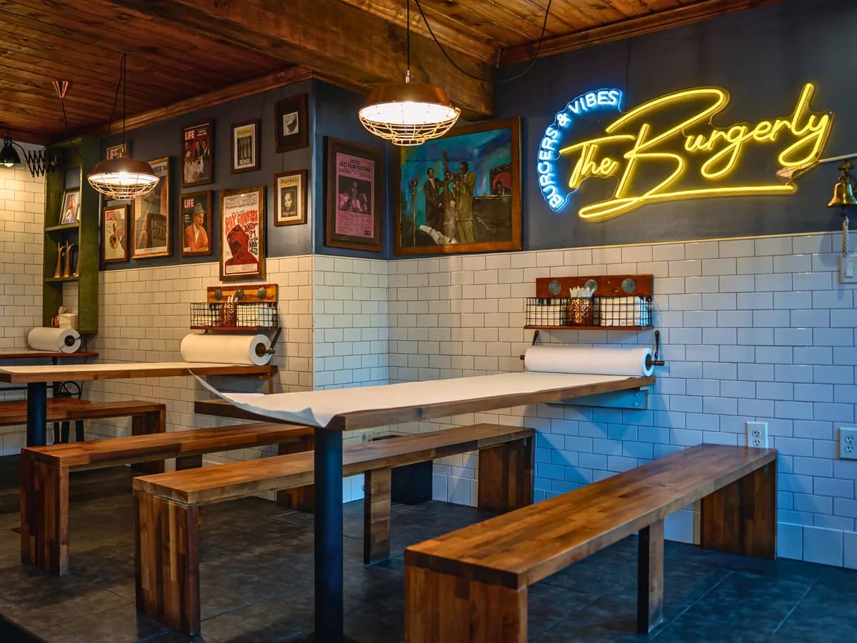 The room at The Burgerly — long communal tables with parchment rolls and benches, under the jazz wall and the yellow neon Burgerly sign.